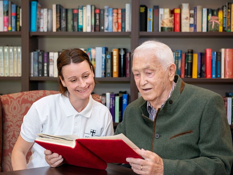 Ein älterer Mann liest aus einem Buch während eine Pflegerin zusieht