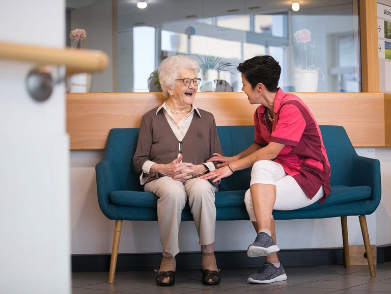 Eine junge Pflegerin und eine Bewohnerin sitzen auf einem Sofa und lachen