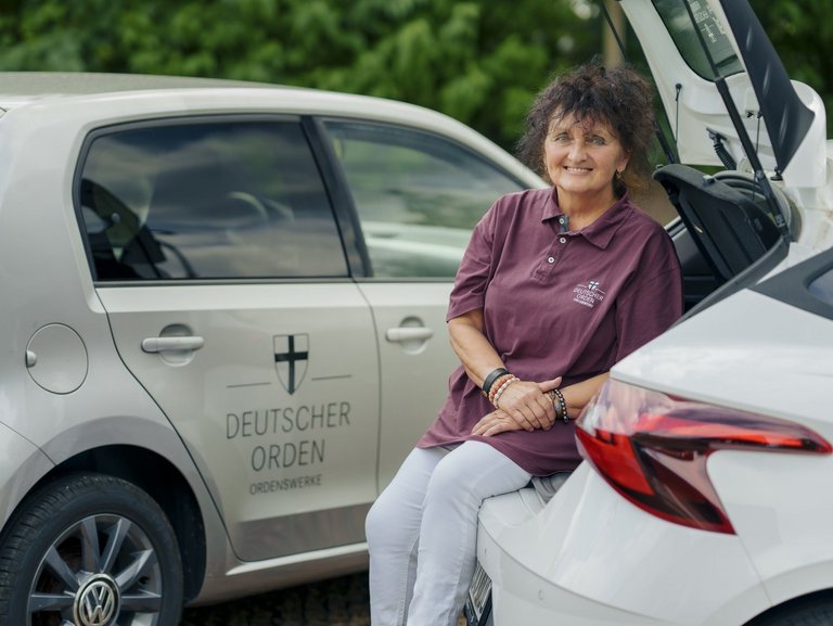 Eine Mitarbeiterin des ambulanten Pflegedienstes lehnt an ihrem Dienstfahrzeug und lächtelt in die Kamera.