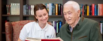 Ein älterer Mann liest aus einem Buch während eine Pflegerin zusieht