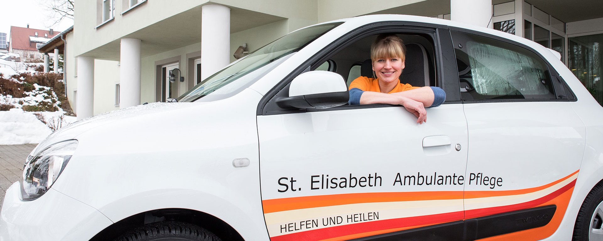 Eine junge Pflegerin in ihrem Auto mit der Aufschrift: "St. Elisabeth Ambulante Pflege - Heilen und Helfen"