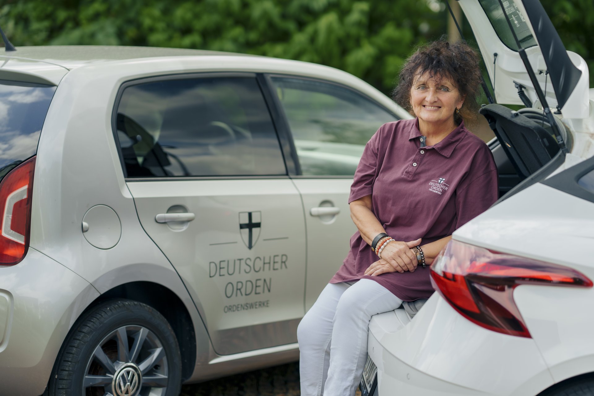 Eine Mitarbeiterin des ambulanten Pflegedienstes lehnt an ihrem Dienstfahrzeug und lächtelt in die Kamera.